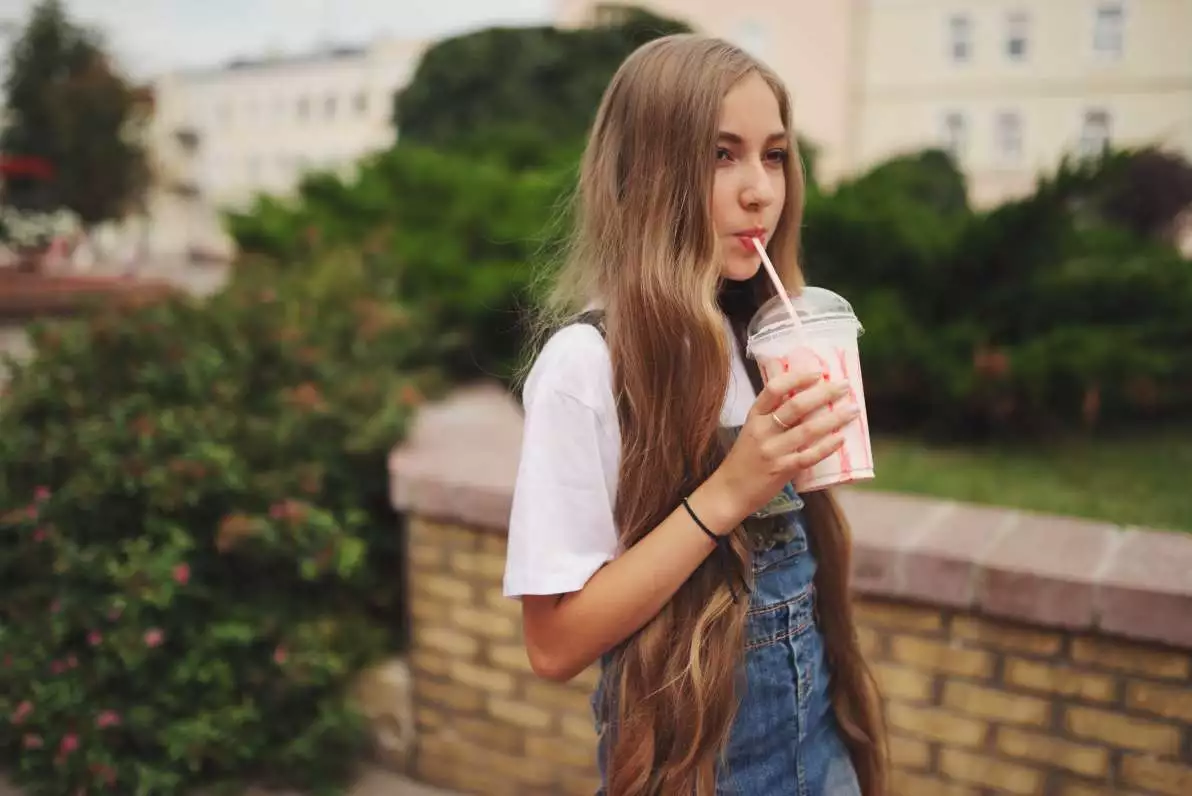 Une jeune fille avec une longue et soyeuse chevelure, en train de boire un smoothie dans un parc.