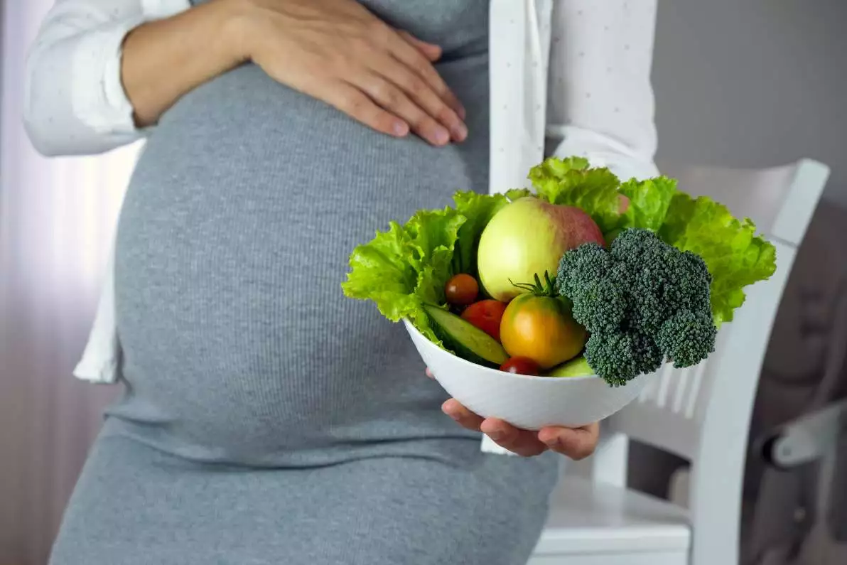 Ventre de femme enceinte avec dans une main une assiette de végétaux, notamment de légumes verts, riches en acide folique.