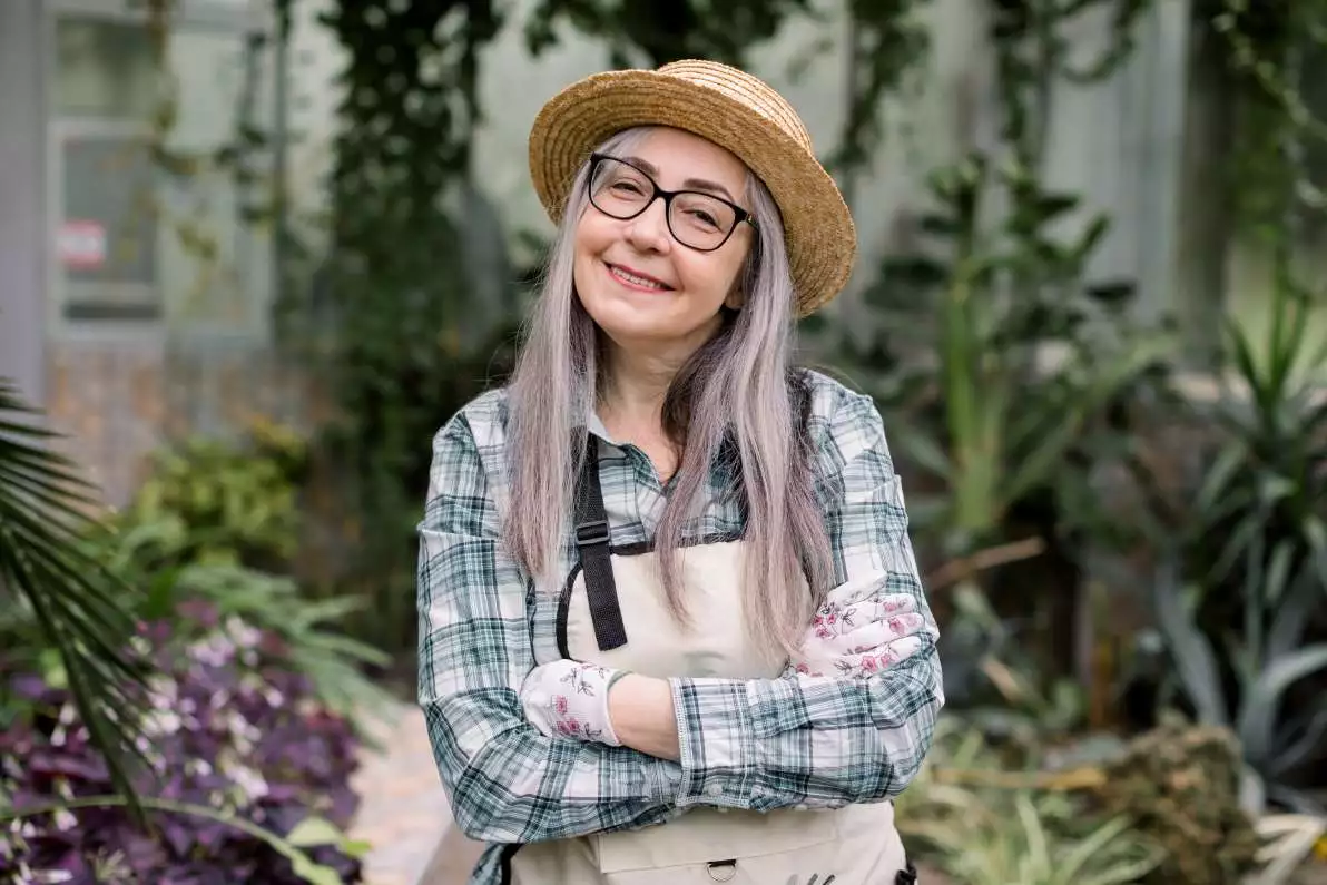 Une dame souriante avec les cheveux grisonnant, se tient debout dans son jardin.