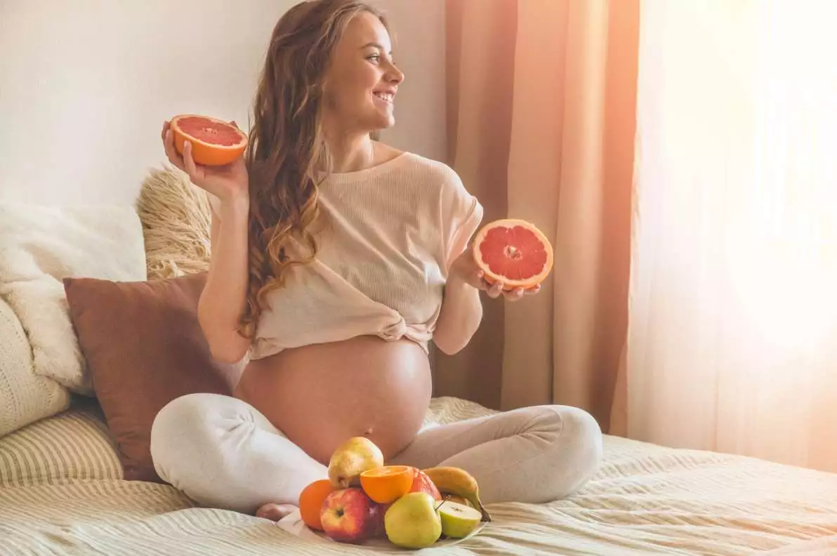 Une femme enceinte est assise sur son lit et tient dans ses mains des végétaux riches en vitamines et minéraux.