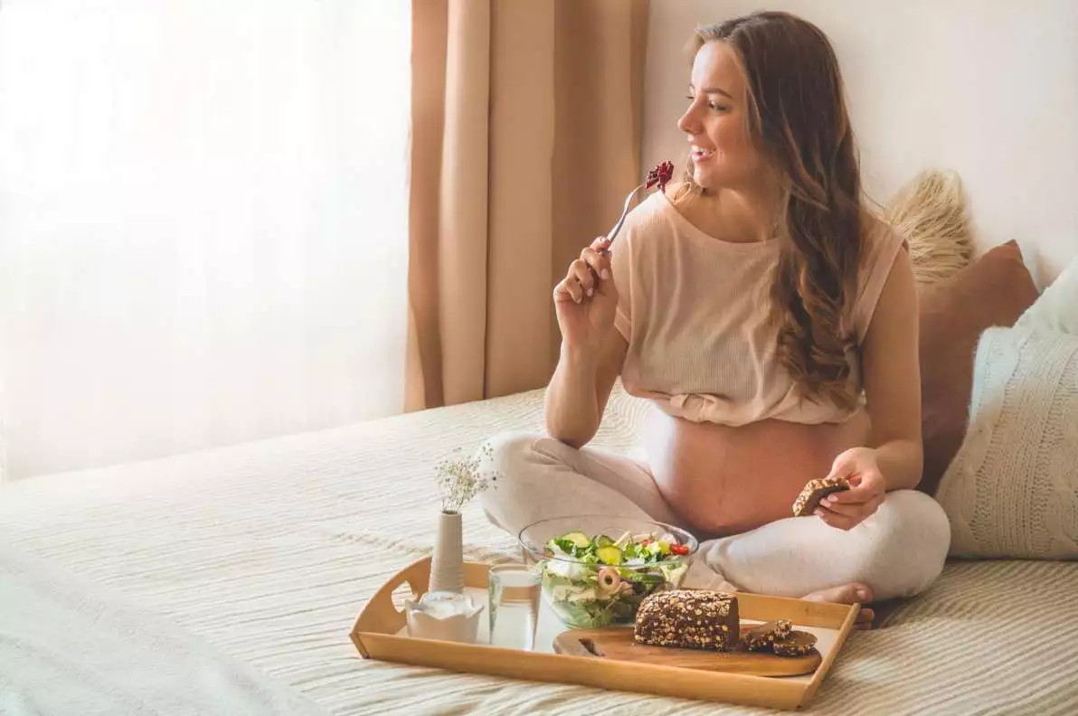 Une femme enceinte est assise sur son lit et mange des végétaux riches en vitamines et minéraux.