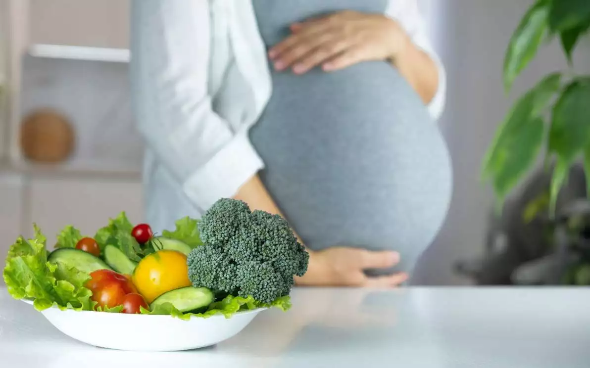 Une femme enceinte en arrière-plan avec une assiette de végétaux riches en acides foliques au premier plan.