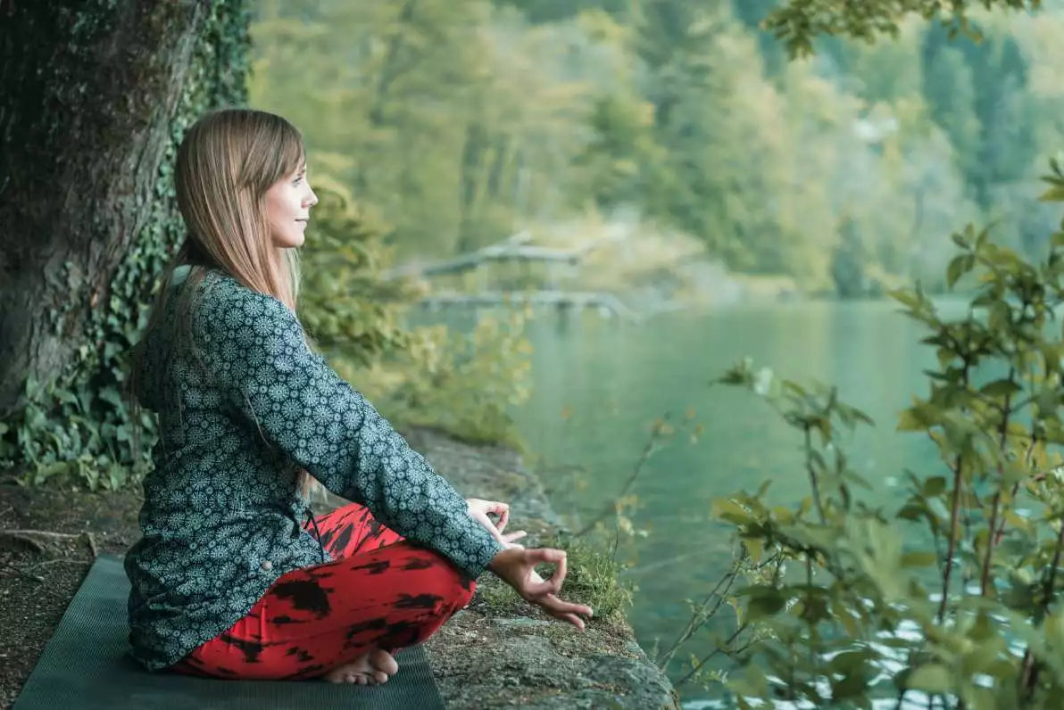 Une femme médite dans la nature, en forêt à côté d’une rivière.