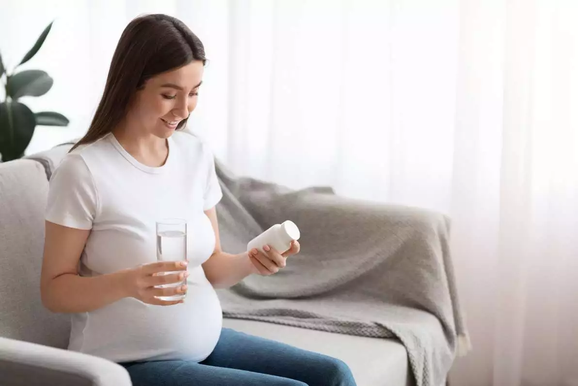 Femme enceinte et souriante, assise sur un canapé et tenant un pot de gélules de vitamines dans une main.