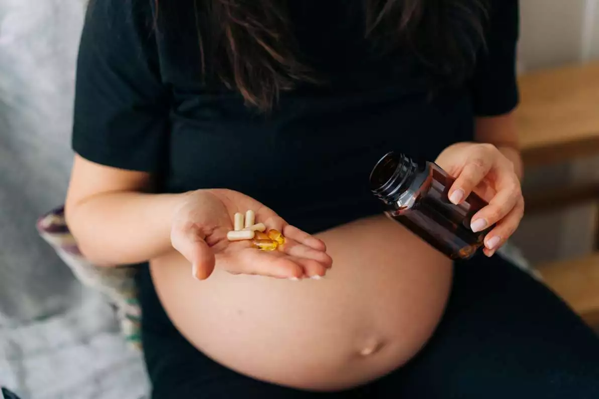 Une femme enceinte est assise sur son canapé et prends des compléments alimentaires vitaminiques.