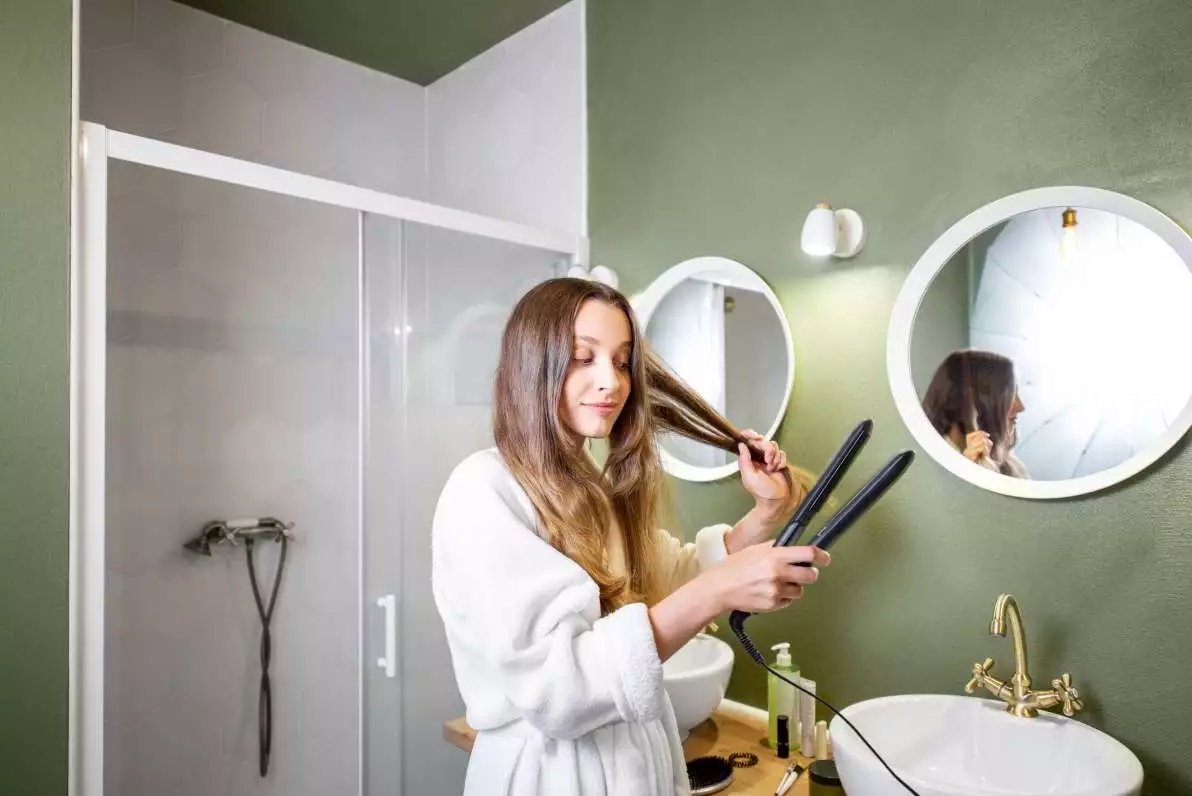 Femme en peignoir se lissant les cheveux dans la salle de bain, à l’aide d’un lisseur éléctrique.