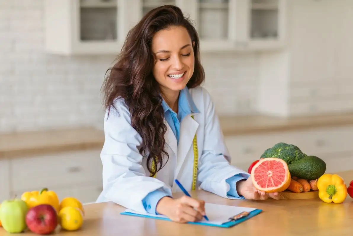 Une nutritionniste souriante en blouse de laboratoire écrit sur un bloc-notes, présentant un assortiment de fruits et légumes frais