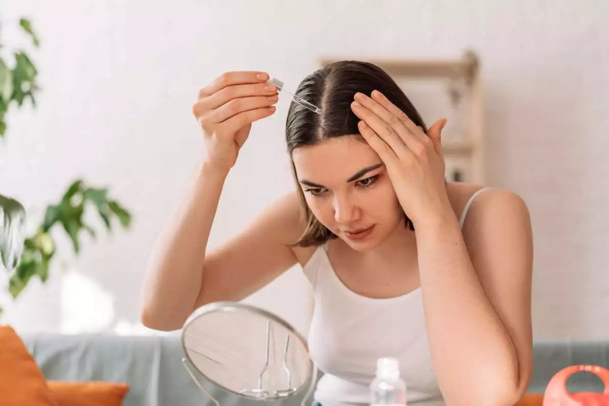 Une dame assise se regarde dans un miroir, tout en se mettant un produit dans ses cheveux, pour favoriser leur pousse.