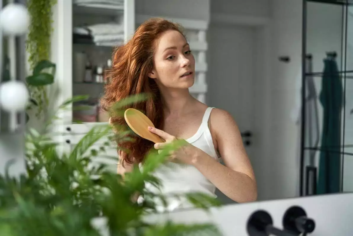 Une femme rousse se brosse les cheveux devant un miroir dans une salle de bain.